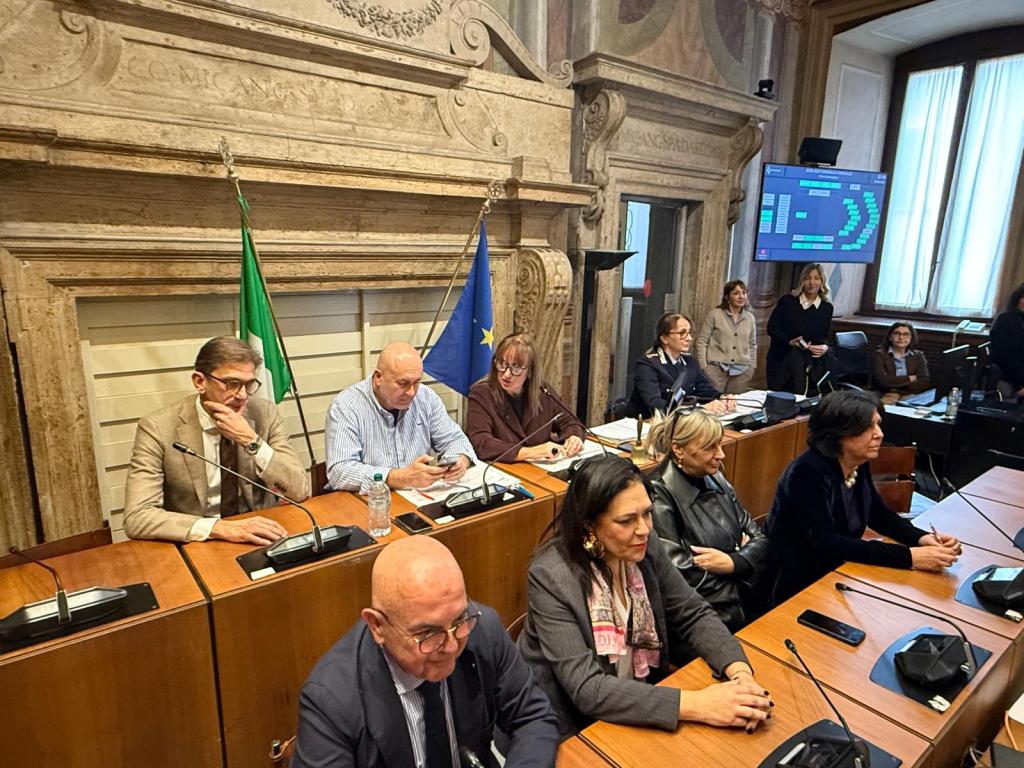 A group of politicians seated at a large wooden table in a formal meeting room, with flags of Italy and the European Union in the background and a display screen showing data.