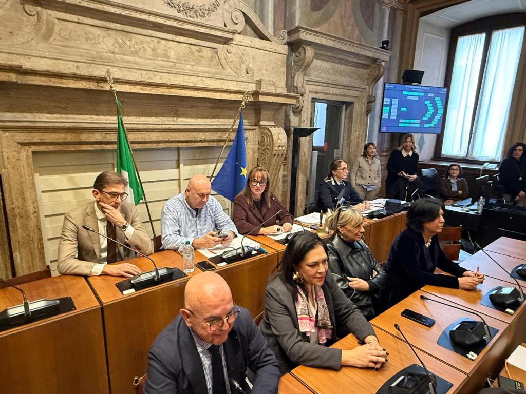 A group of individuals seated at a conference table in a formal meeting room, with flags in the background and a screen displaying data. Some participants are engaged in conversation while others are using their phones.