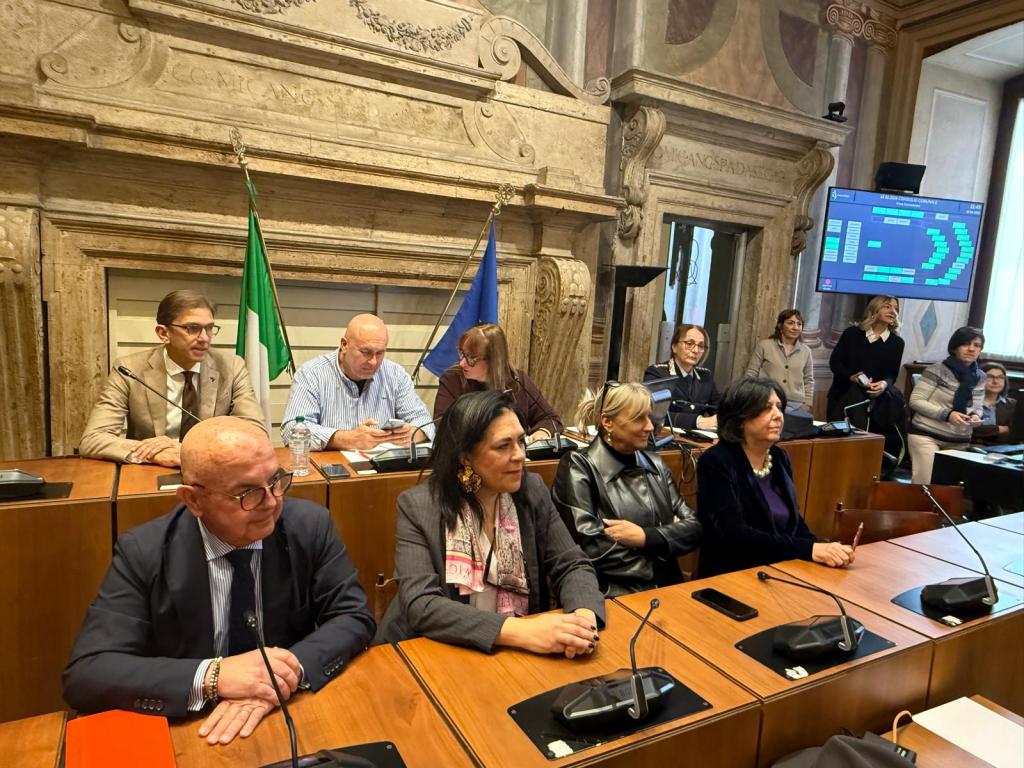 A group of individuals seated at a table in a formal setting, with flags in the background and digital screens displaying information, suggesting a meeting or conference.
