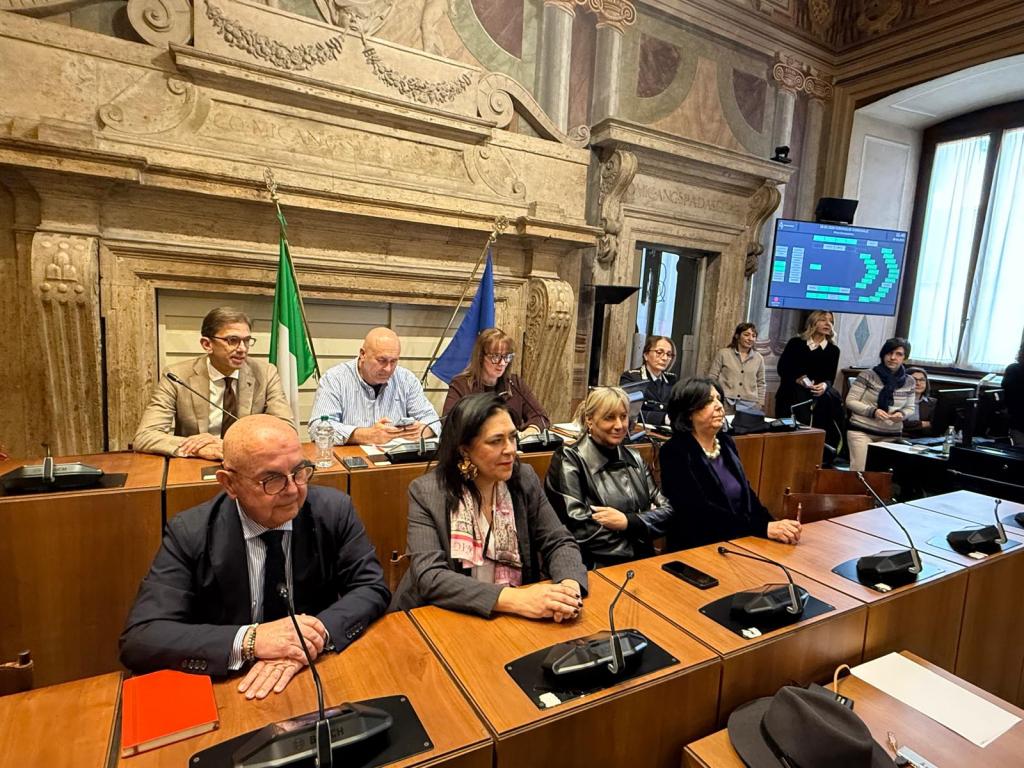 A group of individuals seated at a conference table in a formal meeting room, with flags displayed and a digital screen in the background.