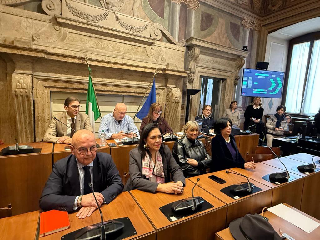 A group of people seated at a conference table in a formal setting, with two Italian flags in the background and a screen displaying information. The individuals appear engaged in discussion or presentation.