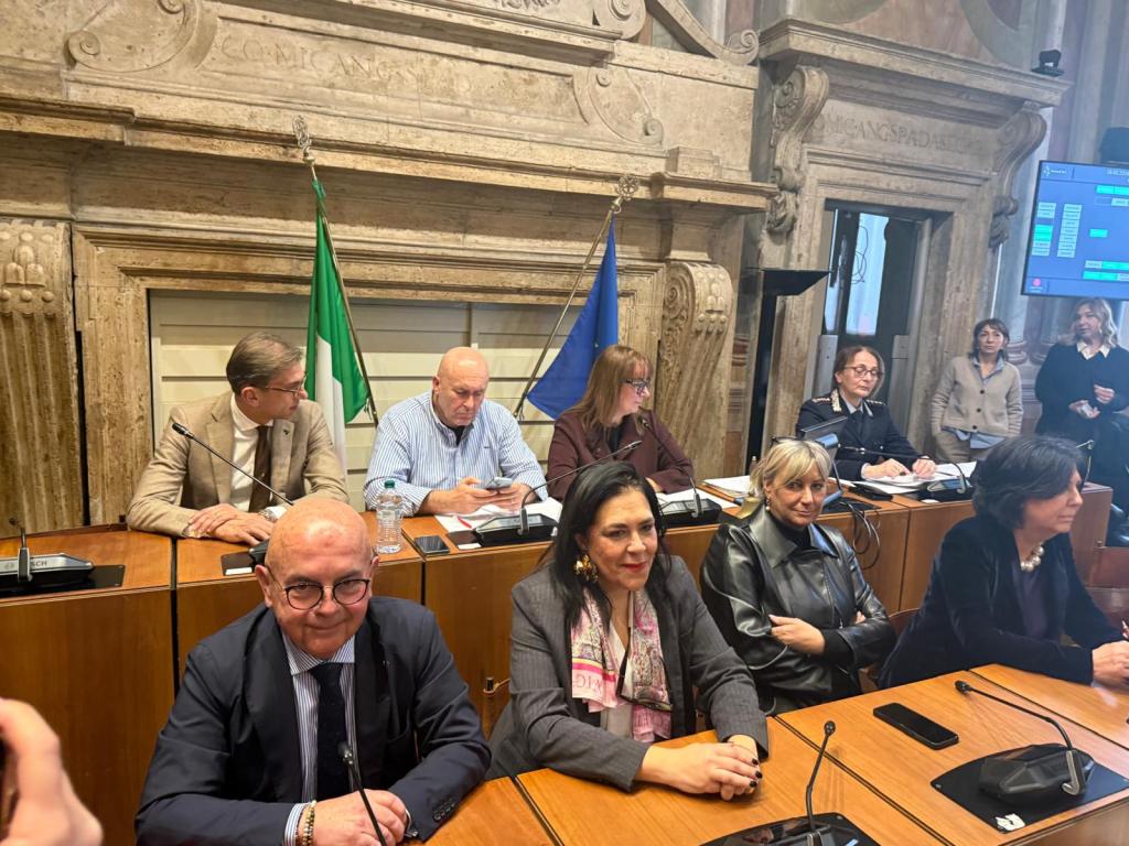 A group of people seated at a formal meeting table, with flags in the background, and a screen displaying information. Some individuals are engaged in discussions while others appear to be taking notes.