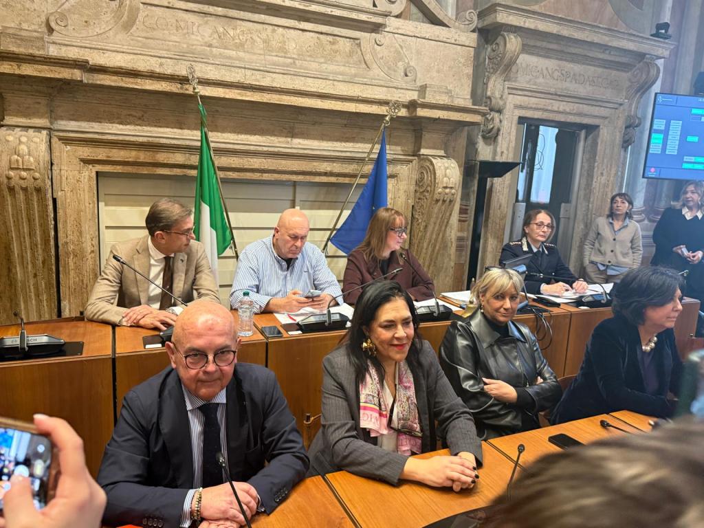 A group of officials seated at a conference table in a meeting room, with Italian and EU flags in the background. Several individuals are engaged in discussion or using electronic devices.