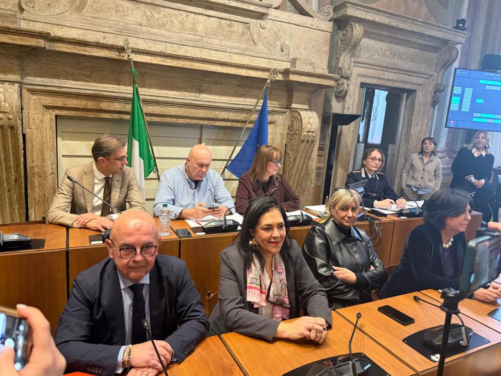 A formal meeting taking place in a governmental setting, featuring several individuals seated at a long table with flags in the background. The group includes men and women engaged in discussion, with some looking at devices or taking notes.