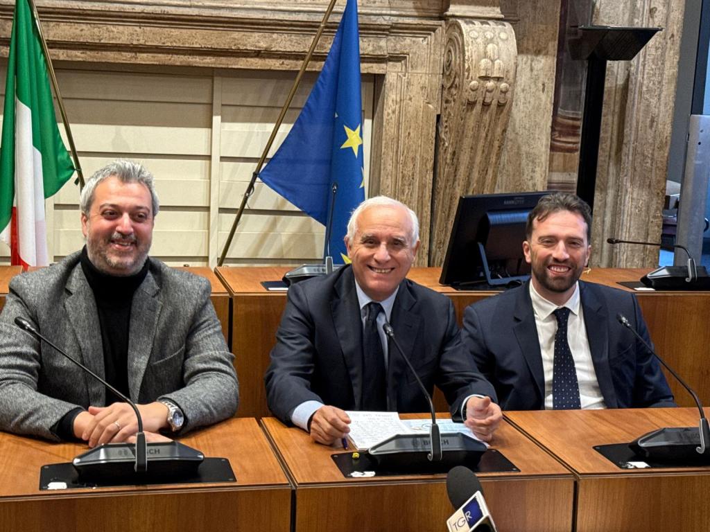 Three men seated at a conference table, smiling and posing for the camera, with an Italian flag and an EU flag in the background.
