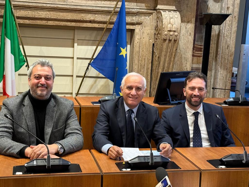 Three men seated at a conference table in a formal setting, with Italian and European Union flags in the background.