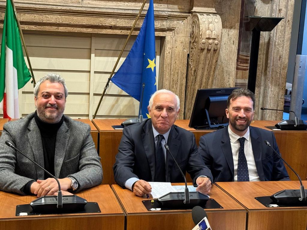 Three men seated at a conference table with national flags in the background.