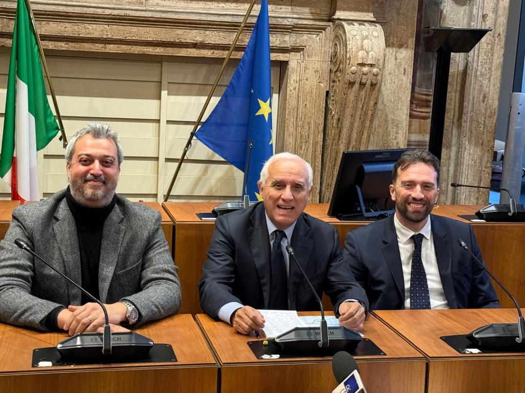 Three men seated at a conference table in front of microphones, with Italian and European Union flags in the background.