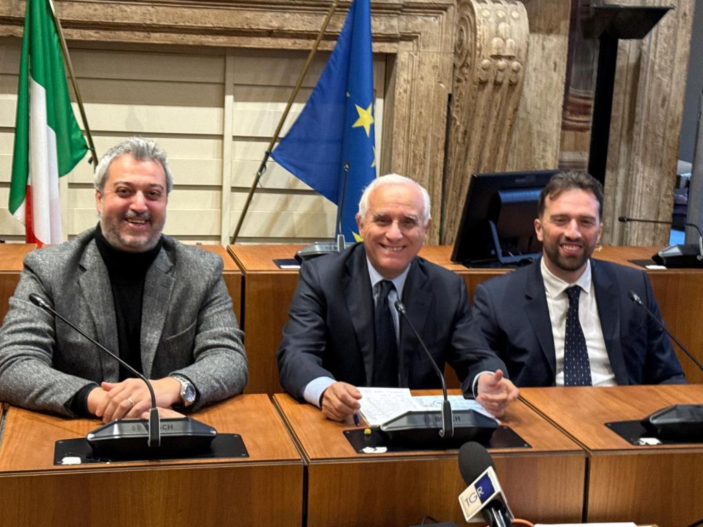 Three officials seated at a table in an official setting, with Italian and European Union flags in the background.