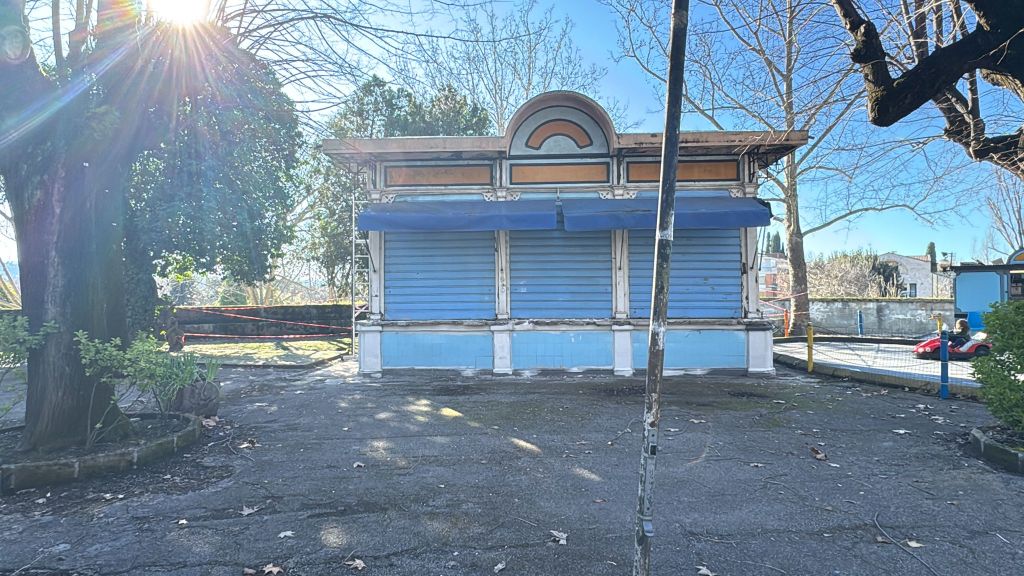 An abandoned blue kiosk with shuttered windows, surrounded by bare trees and overgrown grass in a forgotten park.