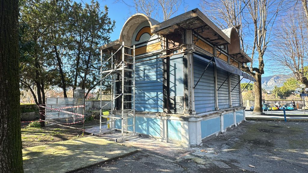An abandoned structure undergoing renovation, featuring scaffolding and closed shutters, surrounded by trees in a sunny setting.