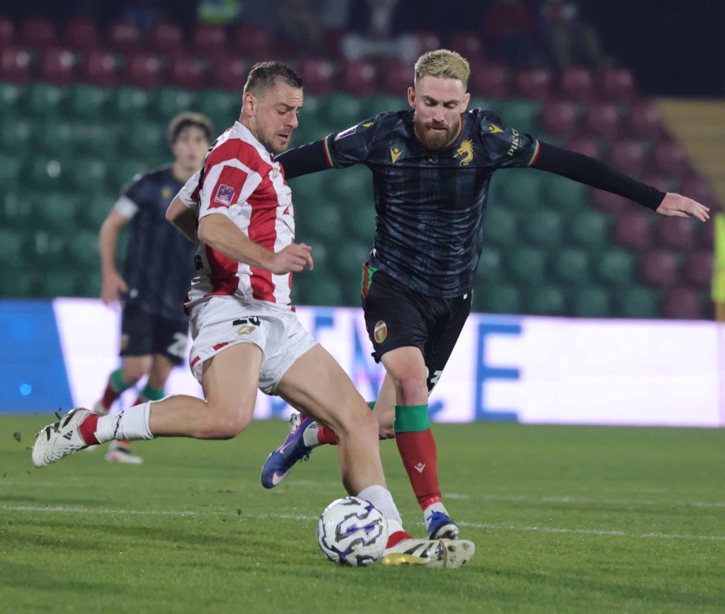 A football match in progress, featuring two players engaged in a competitive play. One player in a red and white striped jersey attempts to control the ball while another player in a black and colorful jersey reaches for it. The stadium audience is visible in the background.