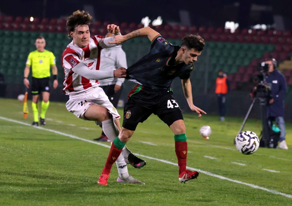 A football match scene showing two players in action, one in a white jersey and the other in a black jersey, competing for the ball. The player in white is pulling the arm of the player in black. An official in a green shirt is visible in the background.