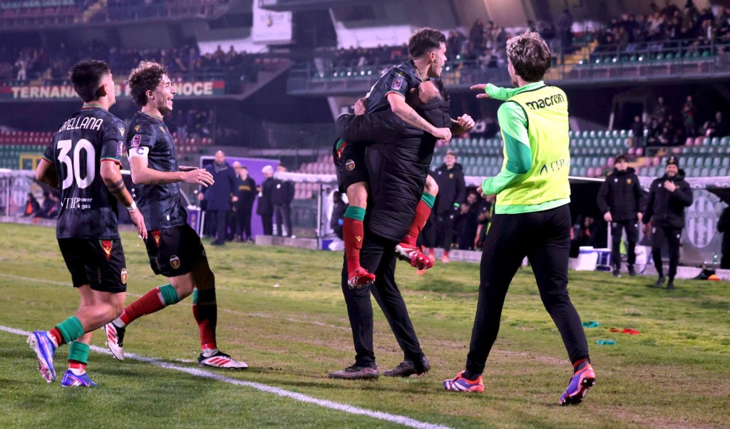A group of football players celebrating on the field after scoring a goal, with one player being lifted by a coach and others joyfully interacting.