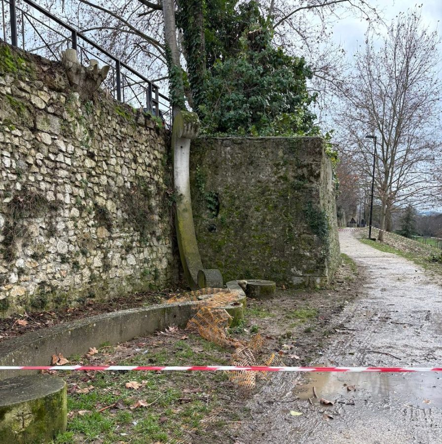 Overgrown stone wall with a concrete structure and an adjacent grassy path, showing signs of wear and neglect.
