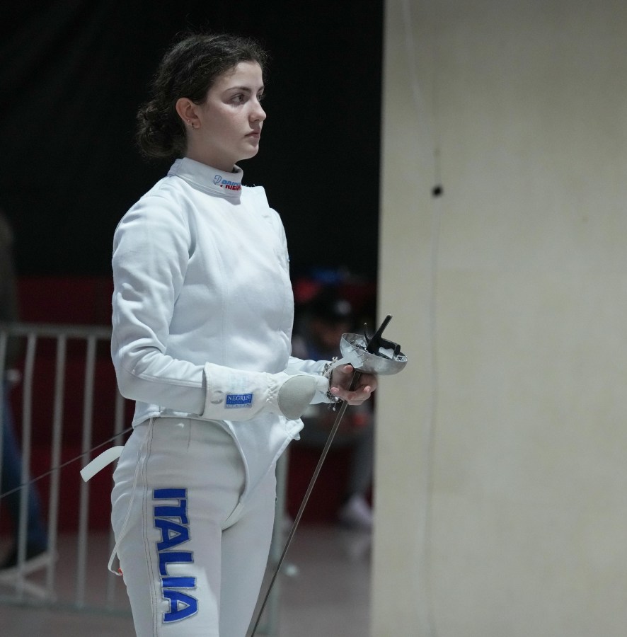 A young female fencer dressed in a white uniform and mask is standing with a fencing sword in hand, focused and ready for action.