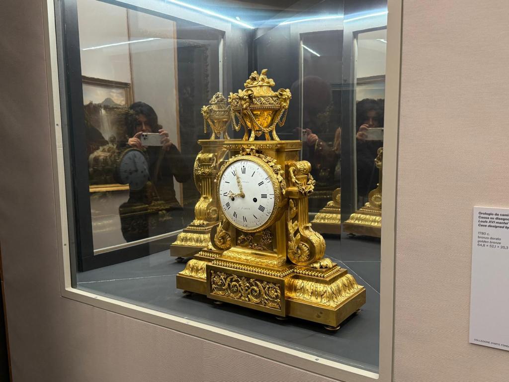 An ornate gold clock displayed in a glass case, featuring intricate designs and a white clock face with black numerals.