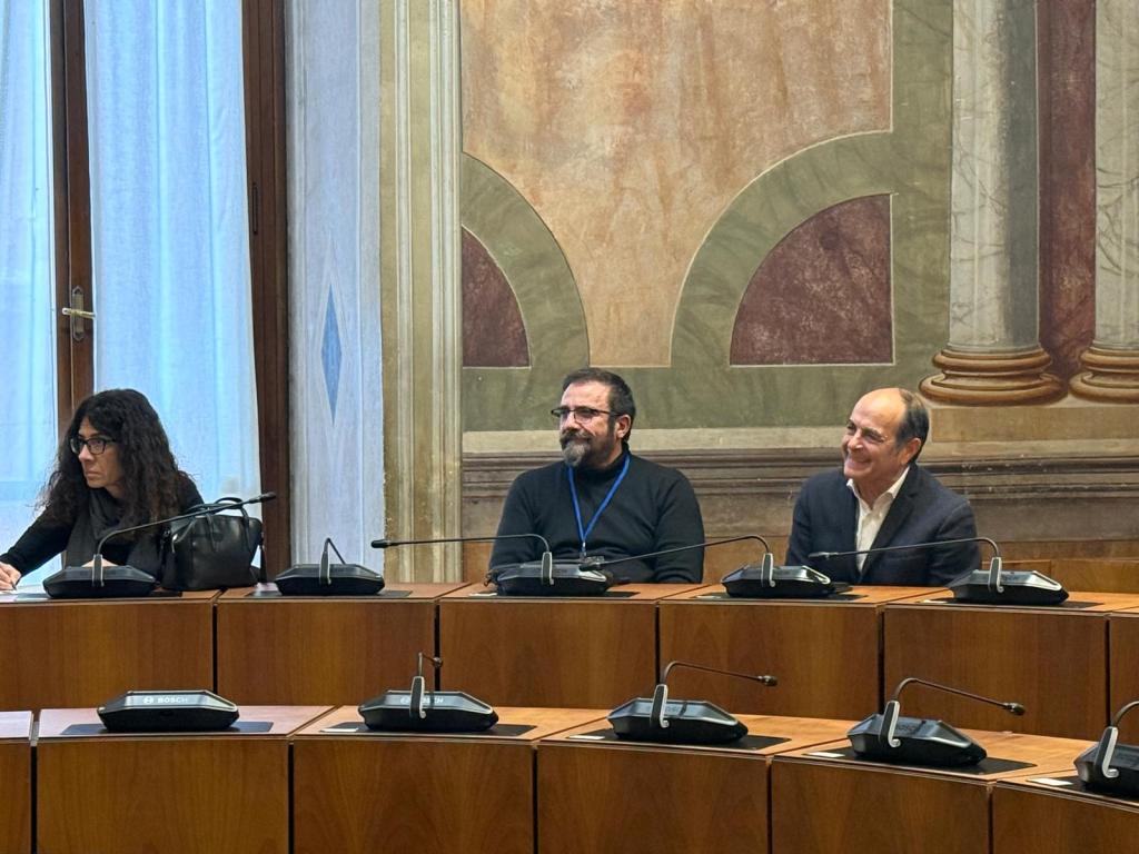 A meeting room setting with three people seated at a table. One person is writing notes, while another is smiling and engaging with the third person. The interior features elegant decor with windows allowing natural light.