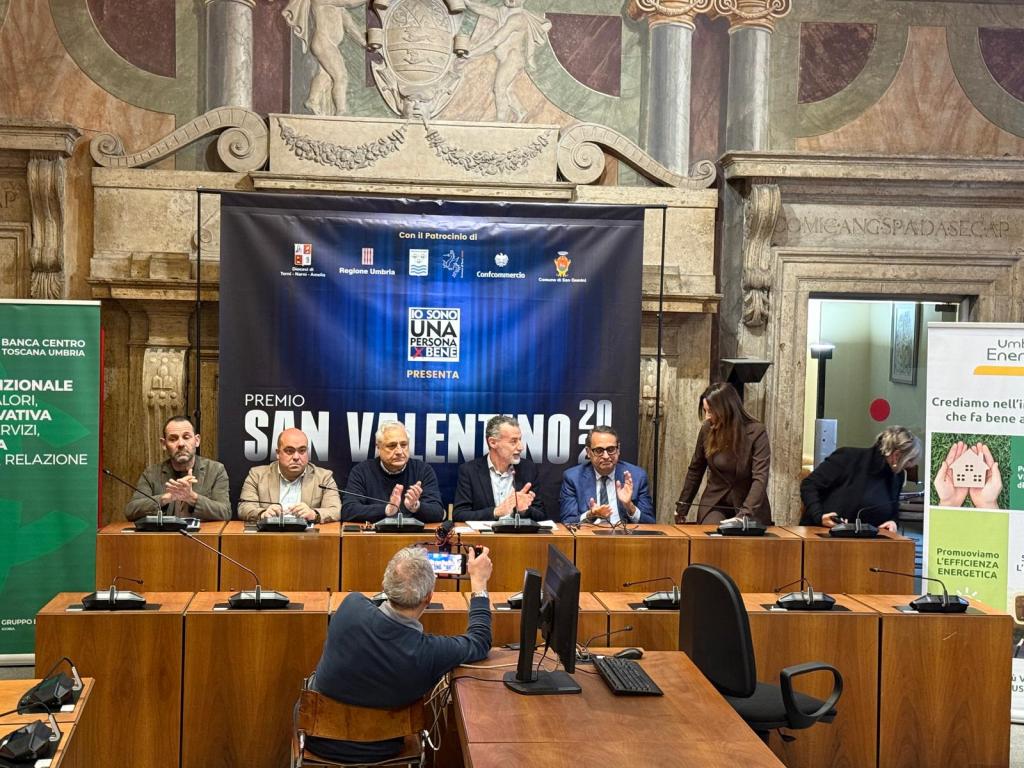 A panel discussion at the San Valentino 2023 award ceremony, featuring several speakers seated at a wooden table with microphones. A large backdrop displays the event title, with logos from various sponsors. Attendees are engaged and taking photographs.