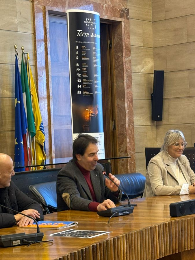 A press conference with three individuals seated at a wooden table, discussing an event. Flags are displayed in the background, and a promotional poster is visible.