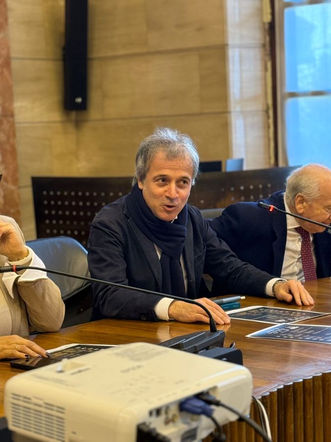 A man in a dark jacket and scarf sits at a conference table, engaging in conversation. Two other individuals are partially visible, one gesturing and the other listening. A projector is in the foreground, and there are documents on the table.