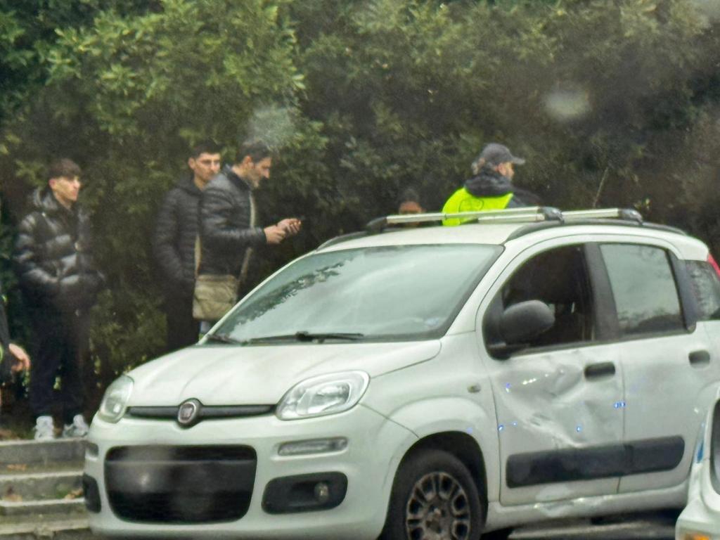 A white Fiat car with a damaged side parked on the street, with several people standing nearby, including a uniformed individual, amid a green leafy background.