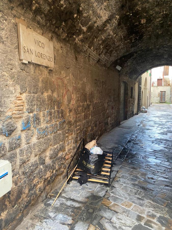 View of a narrow alley with a stone wall, featuring a sign that reads 'Vico San Lorenzo'. A makeshift bed frame with trash bags is on the ground, with a cobblestone path leading to an opening at the end of the alley.