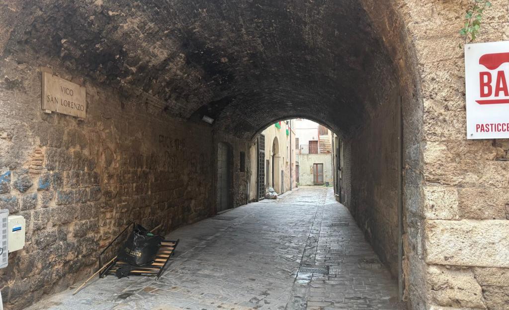 A narrow stone alleyway under an arch with a sign reading 'VICO SAN LORENZO' on the left, leading to a building entrance at the far end. The walls are rustic and weathered, with a red sign for a pasticceria on the right.