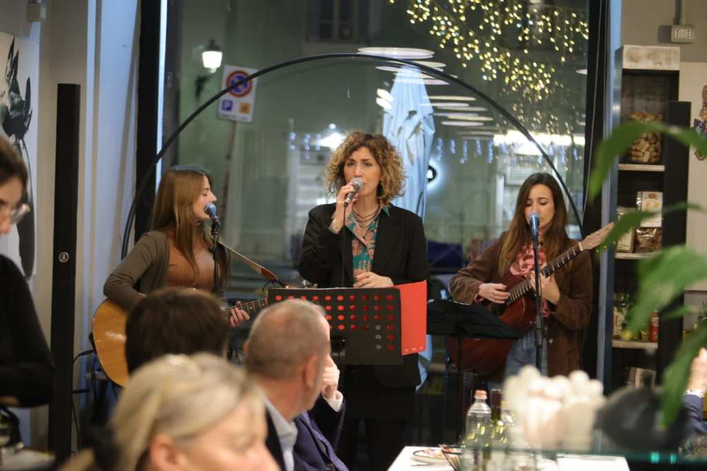 A three-woman musical group performing in a cafe; one woman sings into a microphone while the others play guitar and a smaller string instrument.