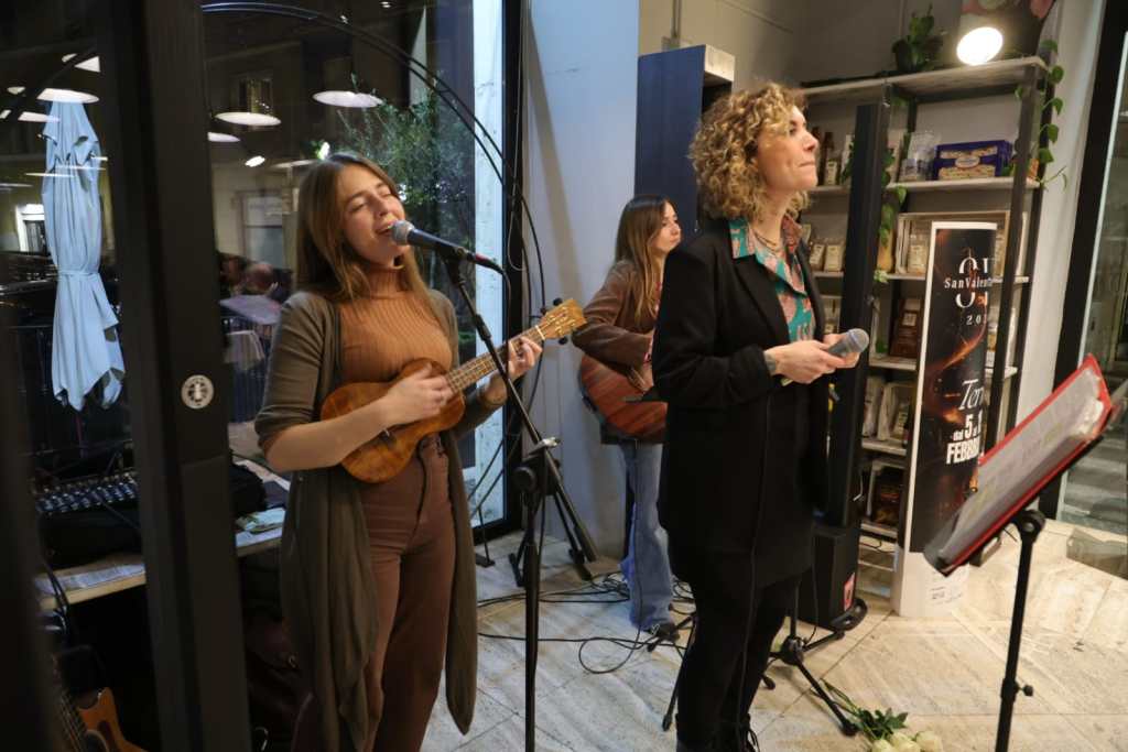 Two female musicians performing in a cozy venue, one singing into a microphone while playing the ukulele, and the other holding a microphone and engaging with the audience.