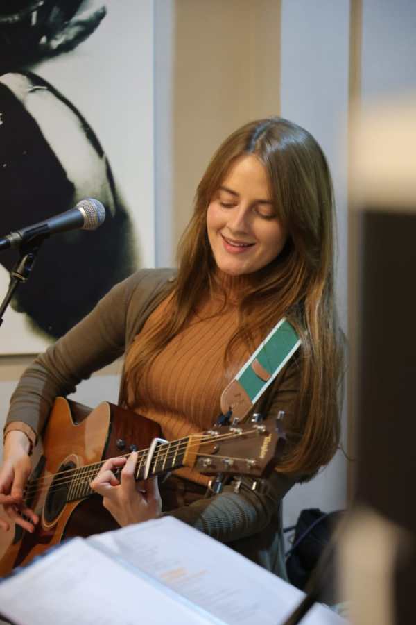 A woman playing an acoustic guitar and singing into a microphone, smiling as she enjoys the performance.