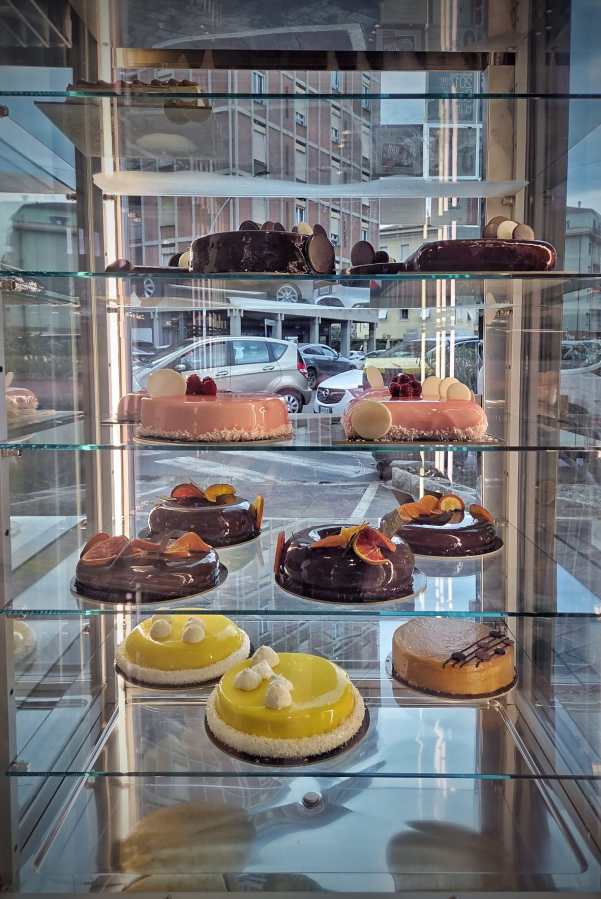 Display case filled with various colorful cakes, including chocolate, pink, and yellow varieties, with decorative toppings like fruits and chocolate pieces.