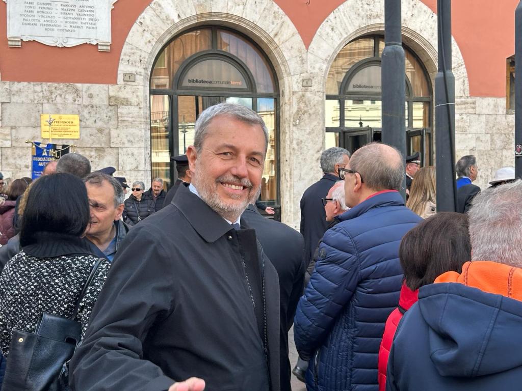 A smiling man in a black coat is standing in a crowd in front of a building with large windows. The background features a stone wall with inscriptions and a sign indicating a library. People are gathered around, creating a lively atmosphere.