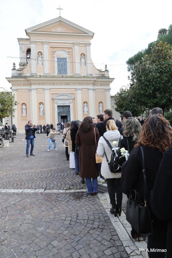A line of people waiting outside a church in an outdoor setting, with a mix of greenery and cobblestone pavement visible.