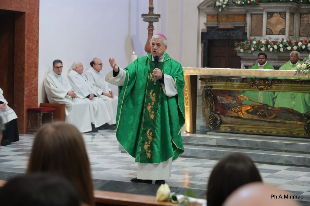 A clergy member in a green vestment, speaking to an audience in a church, with several seated priests and an altar in the background.