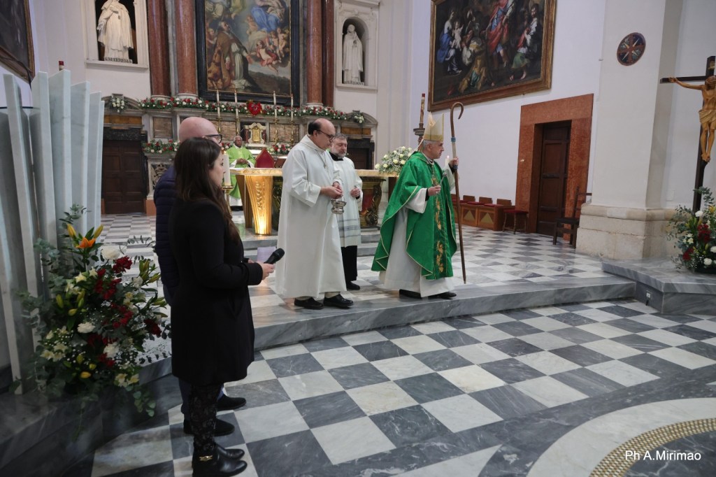 A ceremony in a church with a bishop in green vestments carrying a staff, flanked by other clergy members and a woman holding a microphone. The interior features religious art, floral arrangements, and a patterned floor.