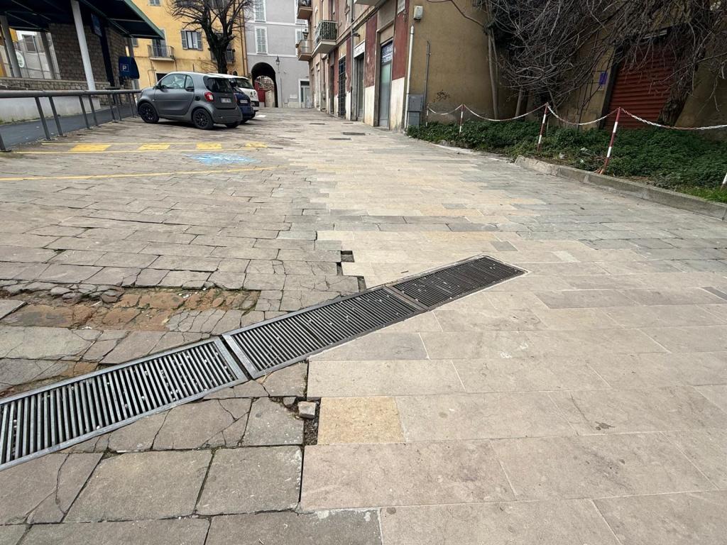 An empty street with uneven cobblestones and a drainage grate, featuring a parked gray car in the background.
