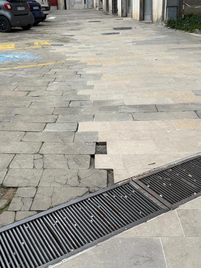 A view of a cracked and uneven pavement with a drainage grate, showing patches of different materials and colors.