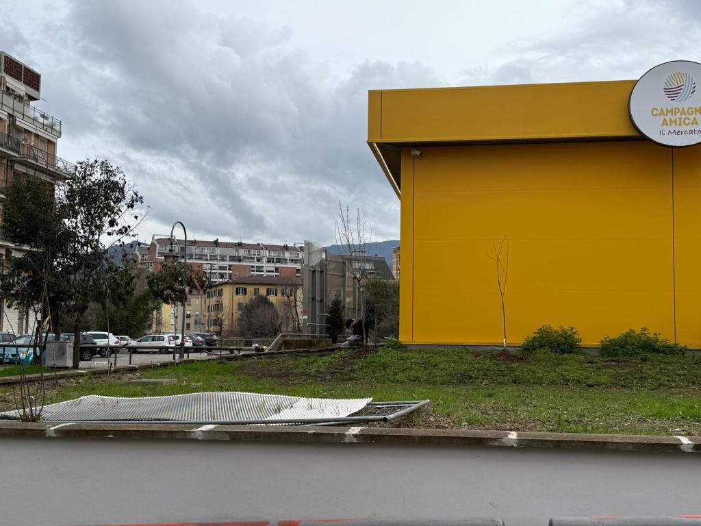 A view of a yellow building with a sign that reads 'CAMPAGNA AMICA' surrounded by green grass and trees, under a cloudy sky, with nearby residential buildings in the background.