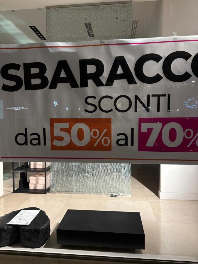 A store window displaying a large banner advertising discounts ranging from 50% to 70%. The banner features bold text with the words 'SBARACCO' and 'SCONTI'.