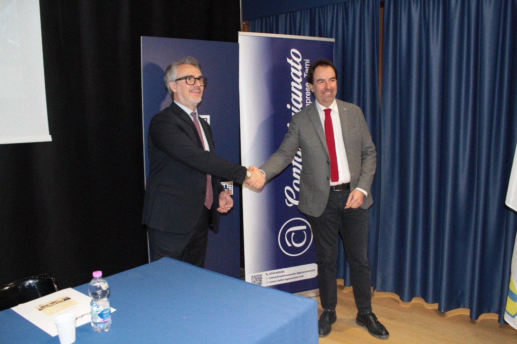 Two men shaking hands at a formal event, with a banner in the background and a table in front.