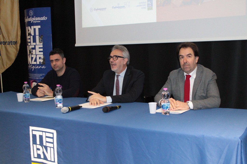 Three men seated at a table during a conference, with water bottles and microphones in front of them, engaged in a discussion.