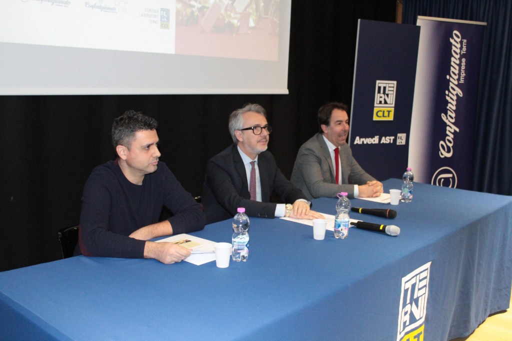 Three speakers seated at a table during an event, with a backdrop featuring organizational logos and a projector screen.
