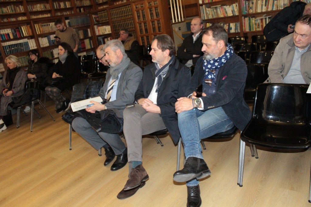 A group of people sitting in a library during an event, some engaged in conversation while others look on attentively, with shelves filled with books in the background.