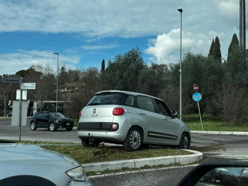 A silver car parked on a grassy area in a roundabout, with trees and cloudy skies in the background.