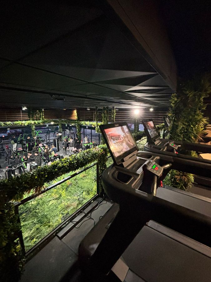 A modern gym interior featuring treadmills with screens overlooking a fitness area filled with people, surrounded by greenery and stylish decor.