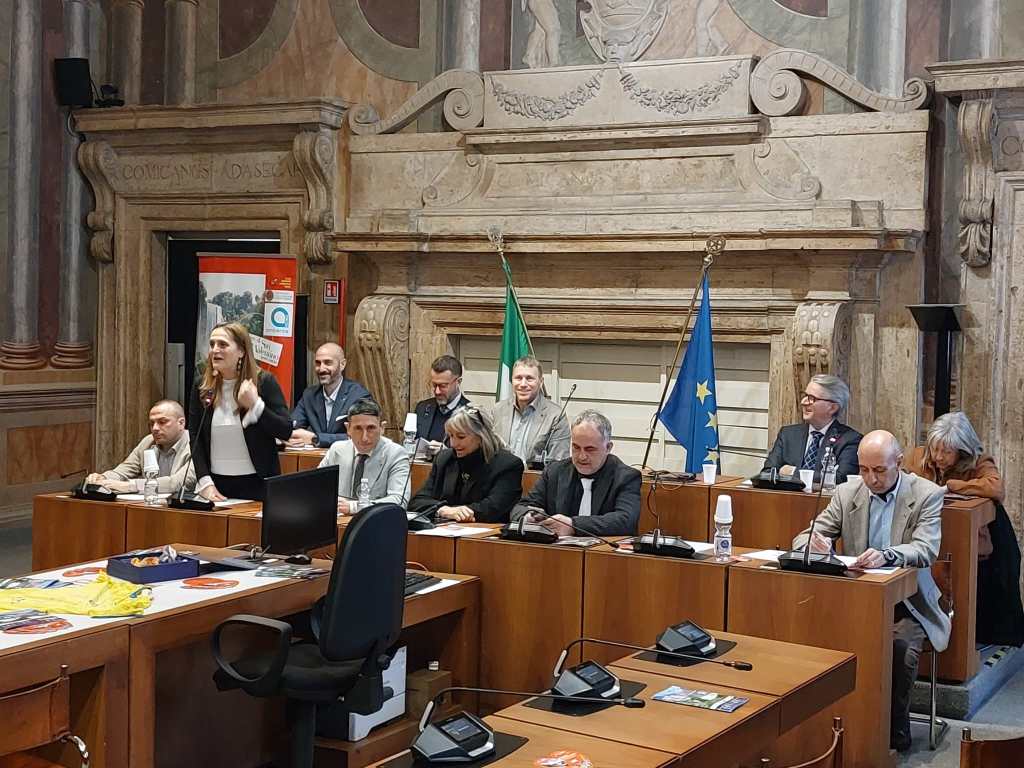 A group of professionals gathered in a formal meeting room with European and Italian flags in the background. A woman is speaking at the front, while others are seated at conference tables, engaged in discussion.