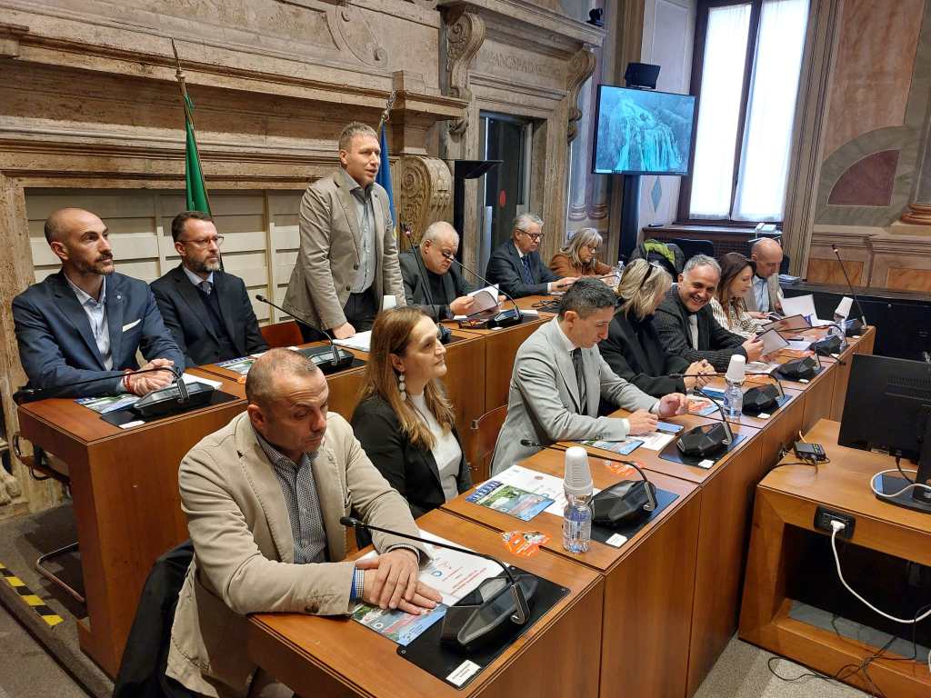A group of people seated at a conference table during a meeting, with some individuals looking at documents and others engaged in conversation. In the background, there is a large screen displaying an image of a waterfall.