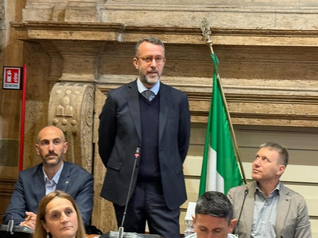 A man in business attire standing while speaking at a table with three seated individuals during a formal meeting, with a green and white flag visible in the background.