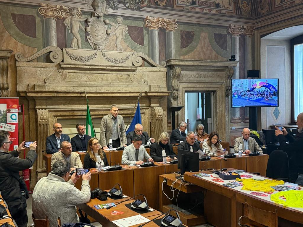 A formal meeting taking place in a grand room with ornate decor. Several individuals are seated at a long table, some taking notes or using their phones. A speaker stands at the front, and flags are displayed behind them. A screen shows images of a colorful event, and various promotional materials are visible on the table.
